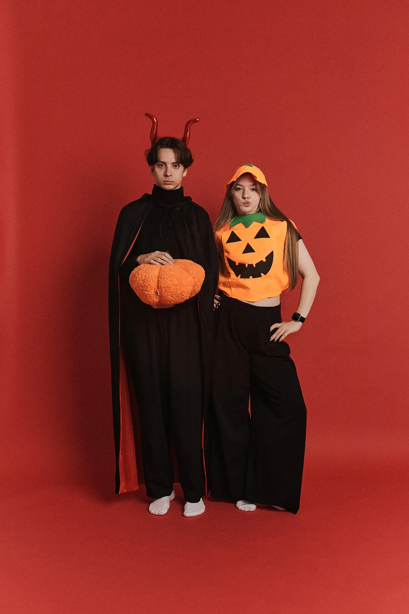 para w halloweenowych kostiumach na czerwonym tle — mężczyzna z rogami trzyma pluszową dynię, kobieta ma strój dyni